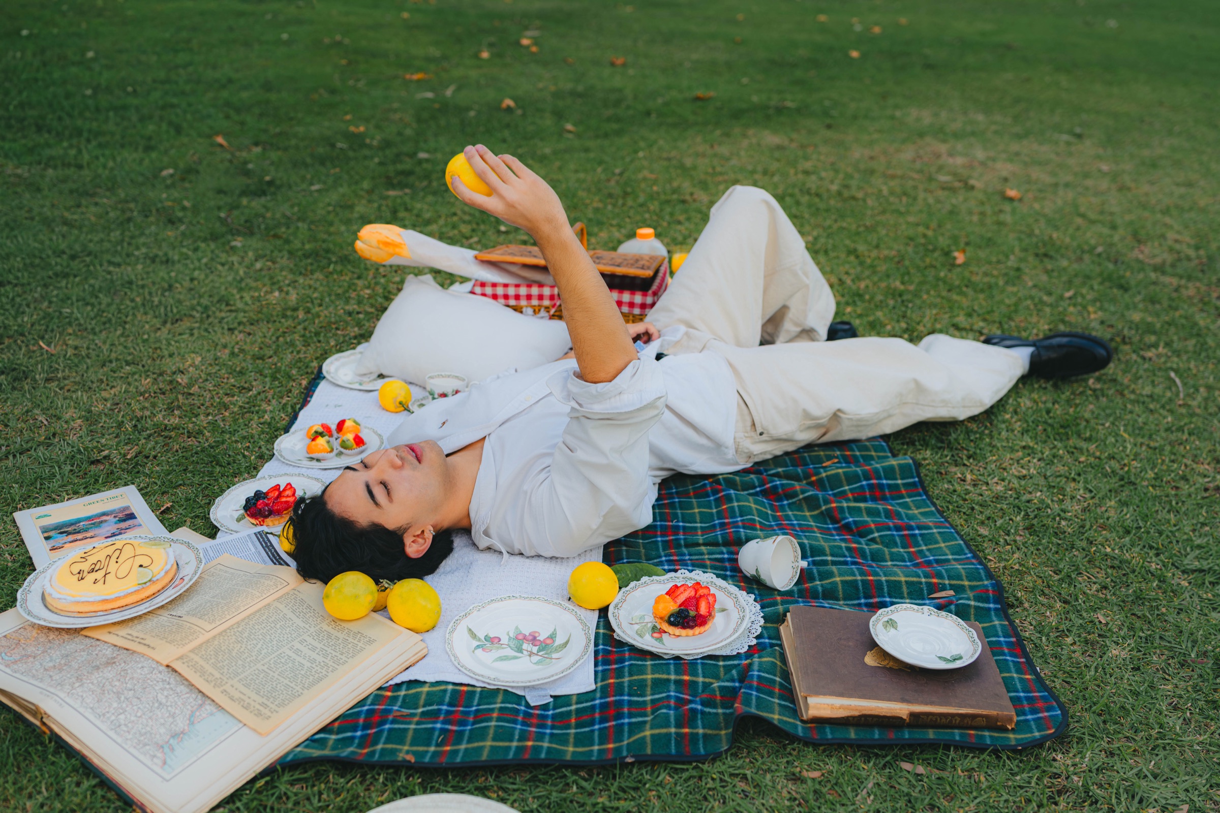 Picnic scene with a model reclining beside books, fruit tarts, and lemons
