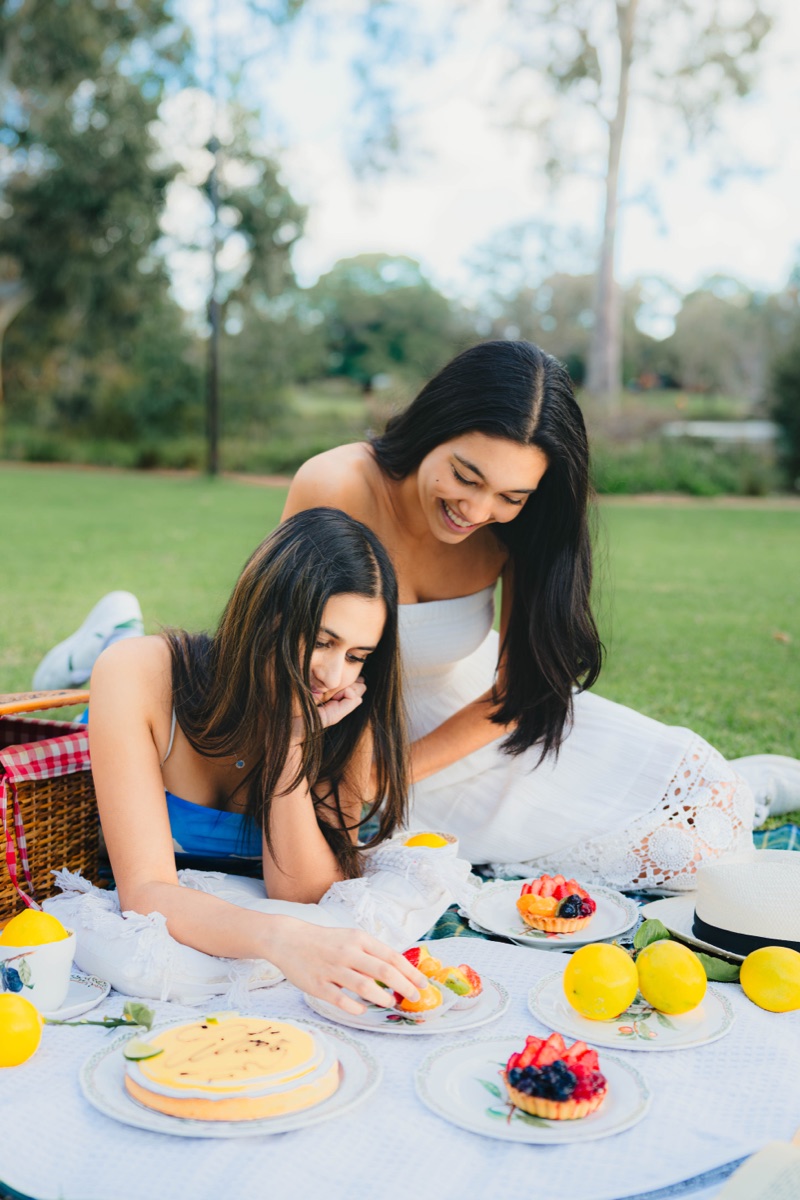Two women at a picnic with lemons, cake, and fruit tarts